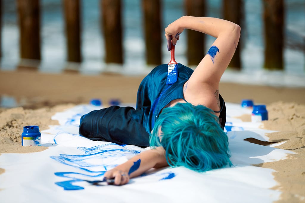 Artistic blue-haired woman performance artist in dark blue dress smeared with indigo gouache painting on her body with brushes on beach. Creative body painting, body art concept, outdoor performance