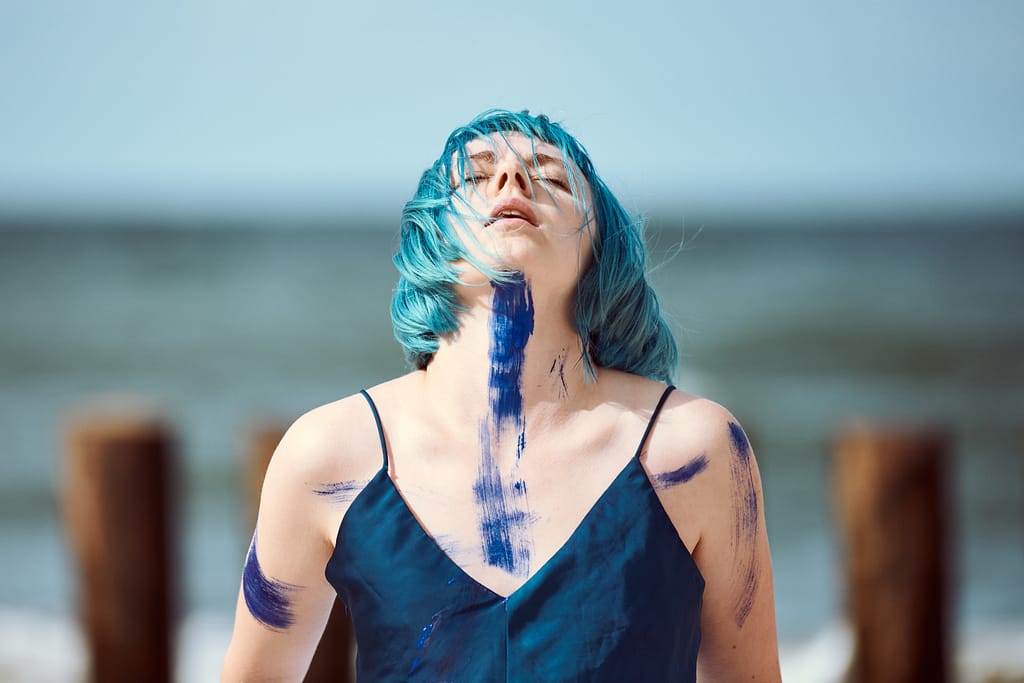 Artistic blue-haired woman performance artist in dark blue dress smeared with blue gouache paints dancing on beach, close up. Body painting and body art concept, expressive performance art outdoor
