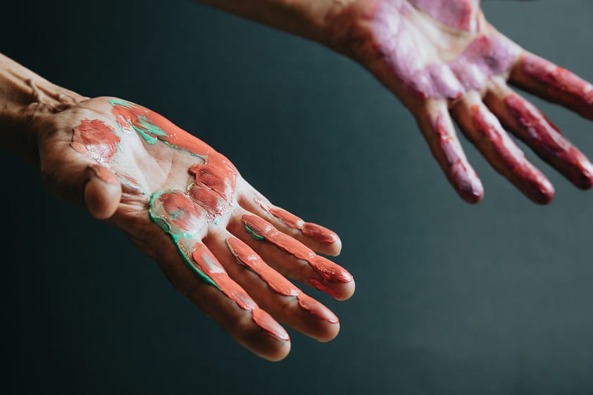 Fun leisure. Creative senior female artist showing hands dirty with red and violet paint. Blur dark background.